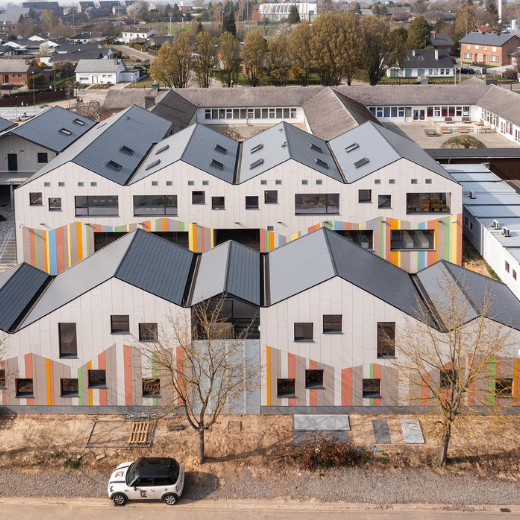 A modern building complex features a series of gabled roofs and a facade decorated with vertical stripes in muted colors. Large windows and sharp lines contribute to its contemporary design. The complex contrasts with the older, traditional buildings in the background, creating a blend of new and classic architecture in a suburban setting.