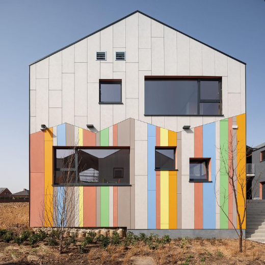 This modern building features a gabled facade with a sleek, neutral gray finish, accented by vertical stripes in earthy tones like orange, green, and blue. Large, asymmetrical windows provide a balance of natural light and visual interest. The colorful stripes create a dynamic contrast against the minimalist upper section, while small windows and ventilation openings add functional detail. The surrounding landscaping is simple, with young plants and a tree complementing the building's clean, contemporary design.