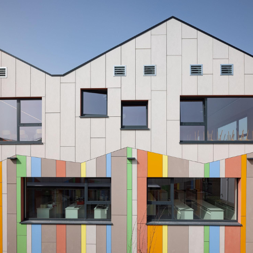 A contemporary building with a dynamic, jagged roofline features an eye-catching exterior design. The facade is a blend of neutral tones accented by vertical stripes in vibrant colors like yellow, orange, green, and blue. Large windows, framed in deep red, allow natural light to flood the interior spaces. The building's upper level includes a series of small square windows, creating a rhythmic pattern across the facade. Below, a recessed entryway is accessed by a set of stairs, adding depth and dimension to the structure. The overall design is playful yet sophisticated, with a focus on clean lines and a harmonious balance of color and form.