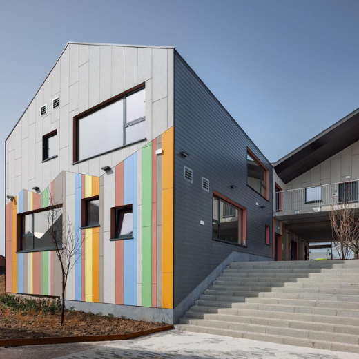 This modern building showcases a clean, geometric design with a gabled roof and a facade featuring vertical stripes in muted tones of orange, green, and blue. The gray exterior contrasts with the colorful accents, creating a balanced, contemporary look. Large windows provide ample natural light, while smaller windows and ventilation openings add functional details. A wide staircase leads up to the building, emphasizing the structure's elevated position. Simple landscaping with young trees complements the sleek architectural lines, enhancing the building's modern aesthetic.