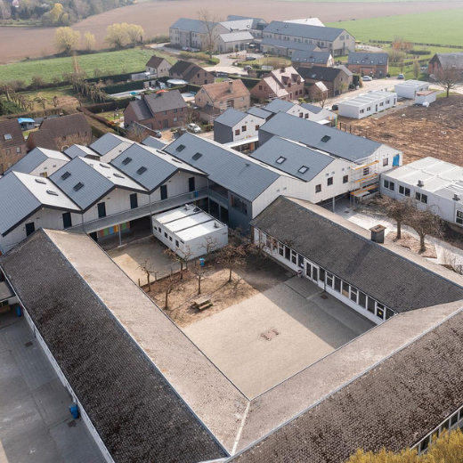 An aerial view of a school complex reveals a blend of old and new architecture. The older, U-shaped building with a central courtyard features a weathered roof and white walls. Behind it, modern gabled structures with sleek gray roofs and clean lines contrast sharply with the traditional design. The complex is set in a rural area, surrounded by additional buildings and open fields.