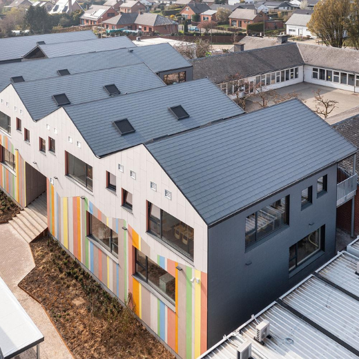 A modern school building with gabled roofs features a vibrant facade of vertical, multicolored stripes. Large windows and clean lines enhance its contemporary design. The structure contrasts with an adjacent older, U-shaped building visible in the background, which has a more traditional appearance with a courtyard at its center. The scene blends modern and classic architectural elements within a suburban setting.